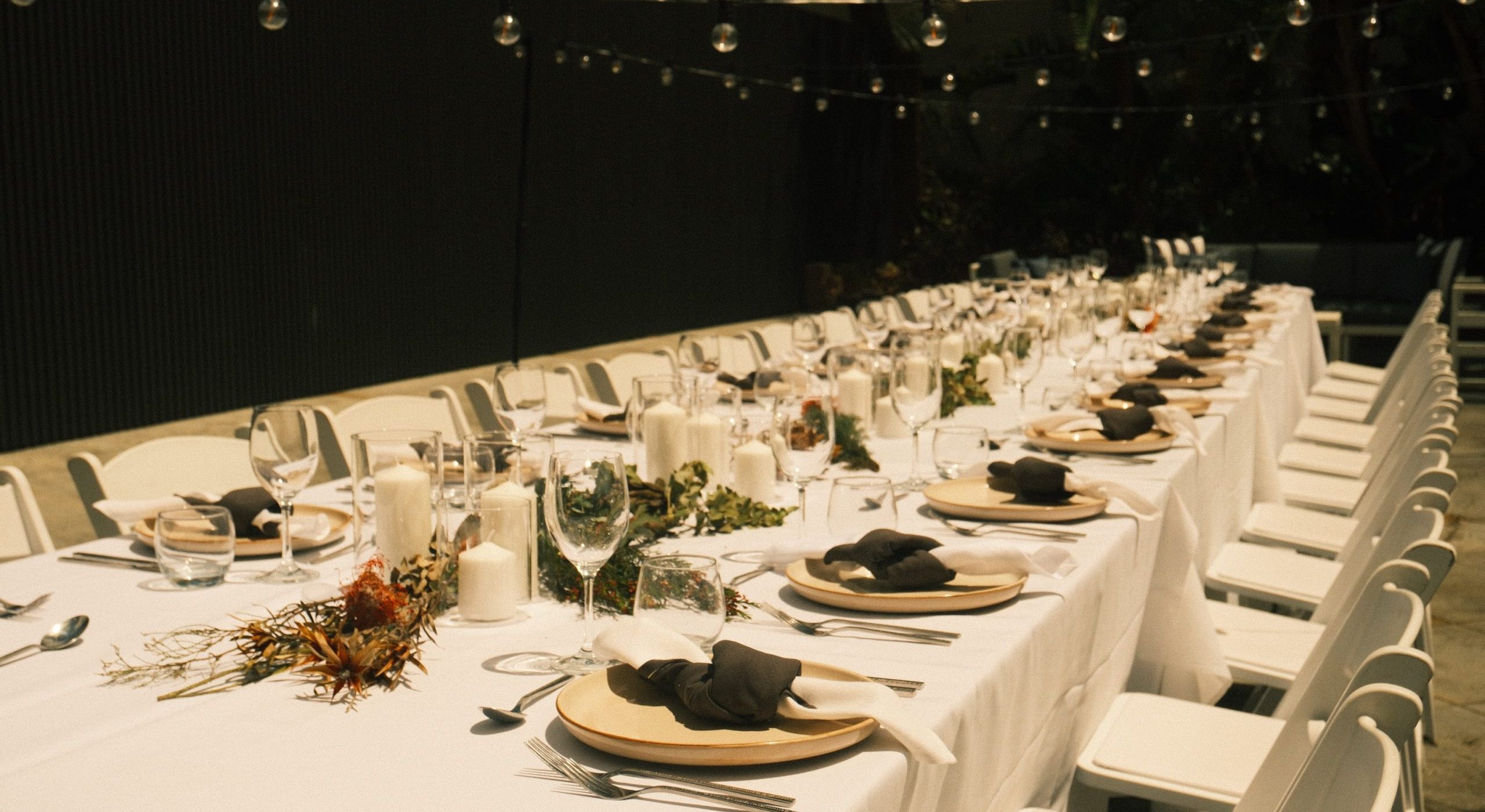 Long outdoor banquet table set with plates, black napkins, glassware, candles, and greenery beneath strings of lights.