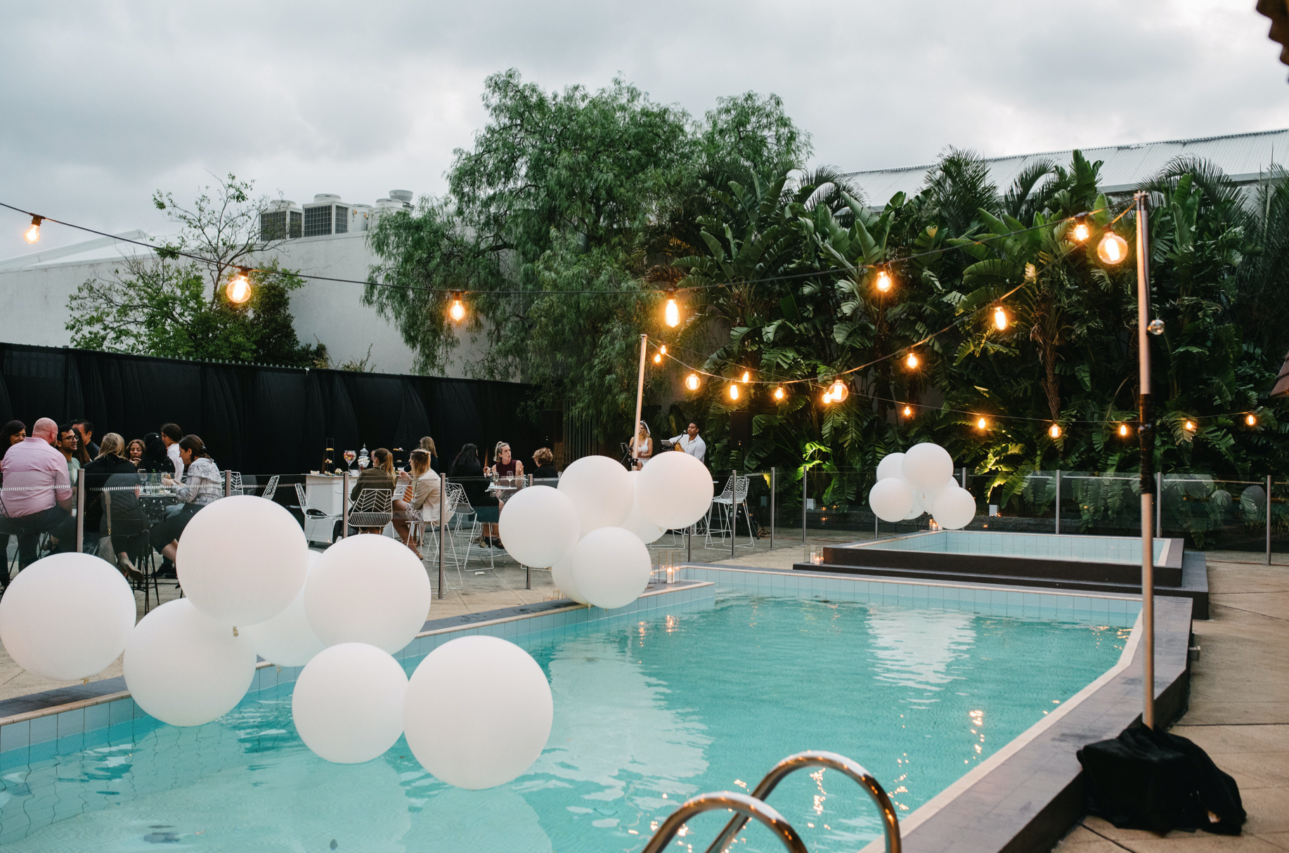 Guests dine beside a turquoise pool decorated with white balloons and glowing string lights, surrounded by tropical greenery.