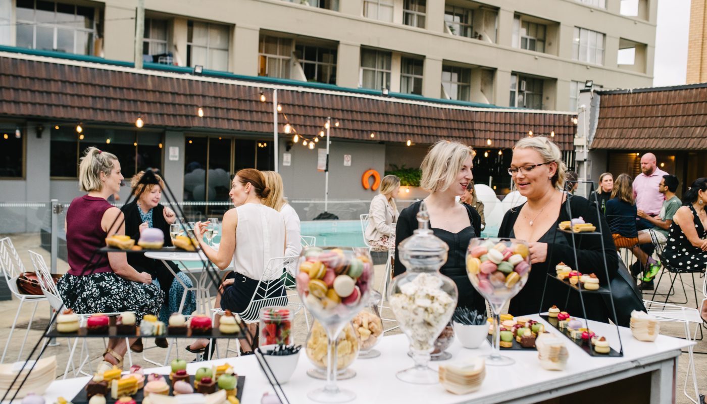 Guests chat at outdoor tables beside a pool, with colorful desserts displayed in the foreground.