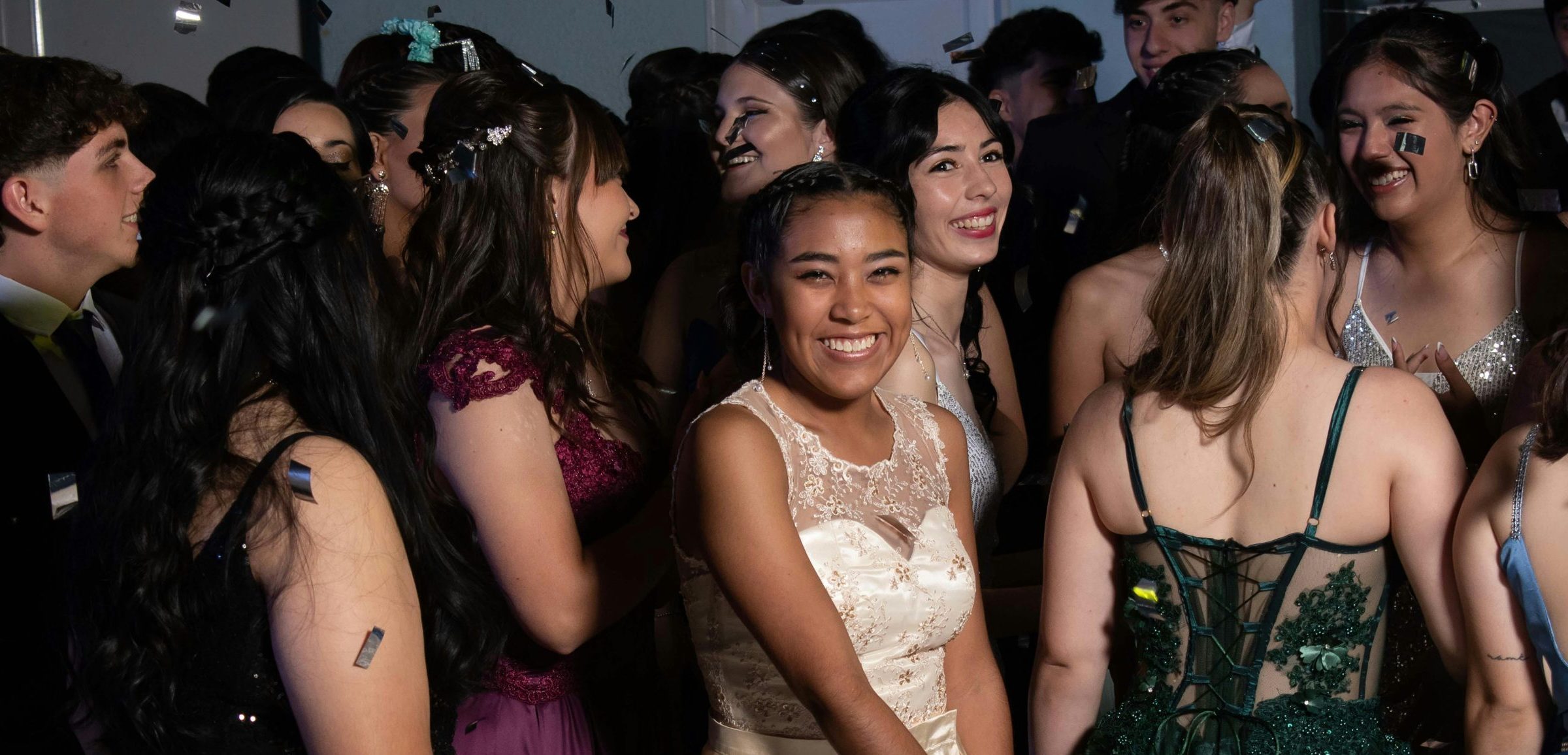 Young people in formal dresses and suits mingle and smile at a crowded celebration, with confetti on their hair and clothing.